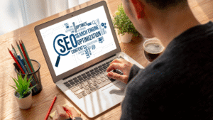 Seorang pria sedang mengerjakan optimasi SEO di laptop dengan tampilan layar bertuliskan “SEO - Search Engine Optimization”, dikelilingi kata kunci digital seperti content, keywords, dan advertising.