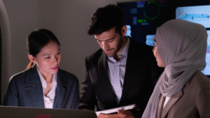 Tiga profesional sedang berdiskusi mengenai strategi digitalisasi dengan layar data analitik di latar belakang.