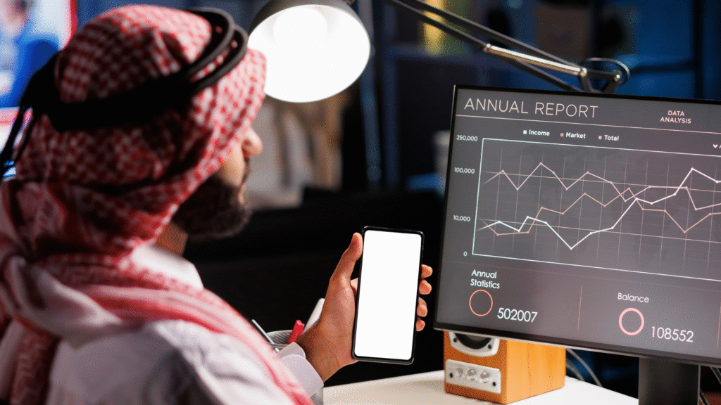 Seorang pria Arab melihat grafik laporan tahunan di layar komputer sambil memegang ponsel kosong, menggambarkan analisis digital untuk strategi marketing biro umroh.