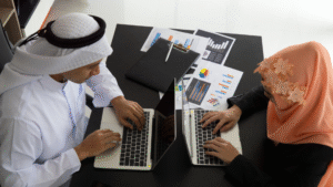 Dua profesional Muslim sedang berdiskusi dan bekerja menggunakan laptop dengan latar belakang grafik data digital, merepresentasikan strategi digital untuk membangun kepercayaan jamaah umroh.