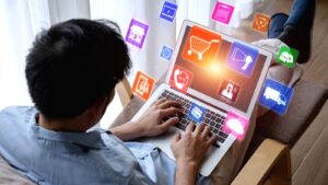 Seorang pria menggunakan laptop dengan ikon digital e-commerce dan layanan online yang muncul di sekeliling layar, merepresentasikan aktivitas belanja online.