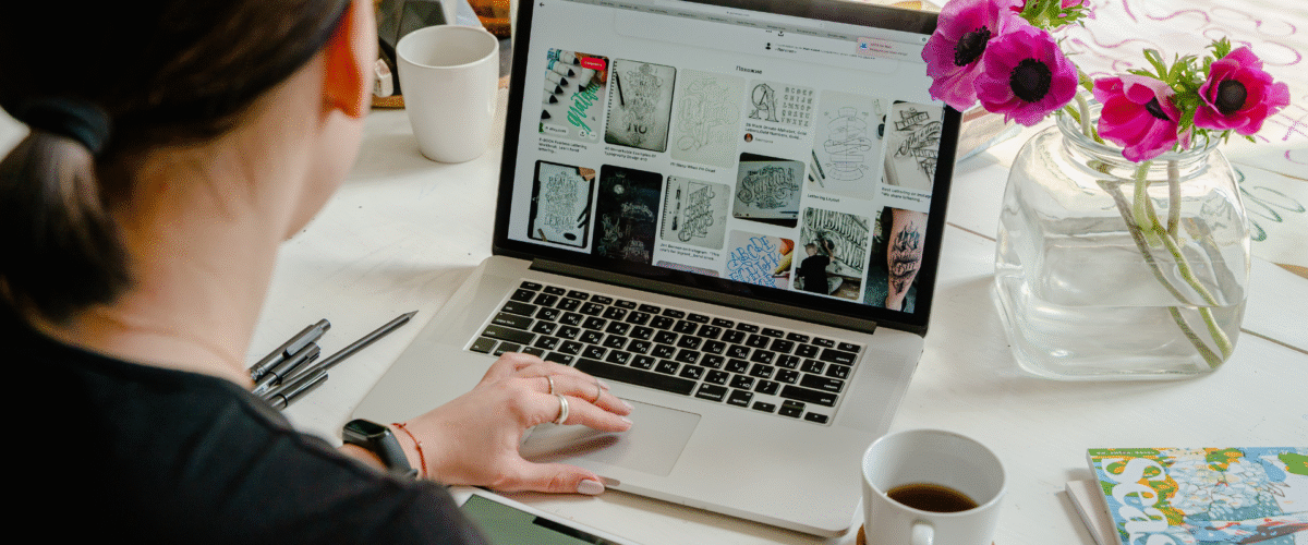 Seorang wanita sedang mencari inspirasi desain visual di Pinterest uuntuk kebutuhan bisnis melalui laptop, sambil memegang tablet bertuliskan “creative”, menunjukkan aktivitas branding digital yang bergantung pada media sosial 