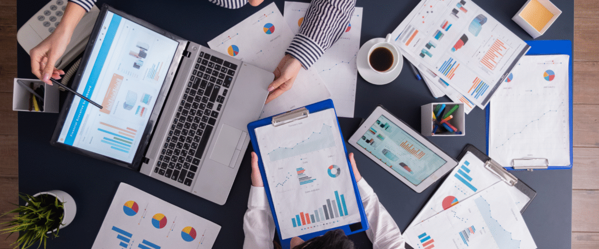 Dua orang sedang menganalisis data grafik dan metrik pemasaran digital di atas meja kerja dengan laptop, tablet, dan dokumen cetak