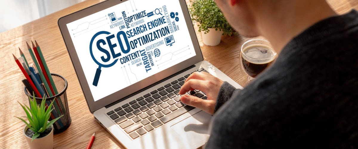 Seorang pria sedang mengerjakan optimasi SEO di laptop dengan tampilan layar bertuliskan “SEO   Search Engine Optimization”, dikelilingi kata kunci digital seperti content, keywords, dan advertising 