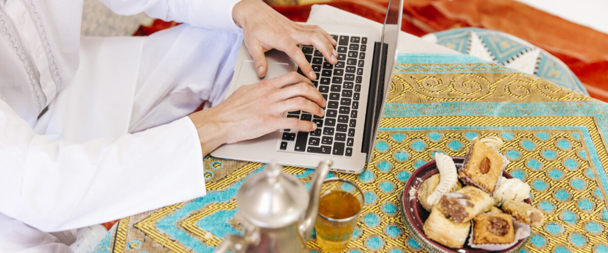 Pria mengenakan baju putih tradisional sedang mengetik di laptop di atas meja dengan taplak bernuansa islami, dikelilingi teh dan kue khas Timur Tengah 