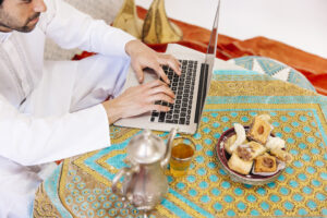 Pria mengenakan baju putih tradisional sedang mengetik di laptop di atas meja dengan taplak bernuansa islami, dikelilingi teh dan kue khas Timur Tengah.