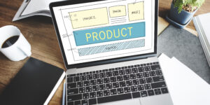 Sketsa desain tampilan website produk pada layar laptop di atas meja kerja.